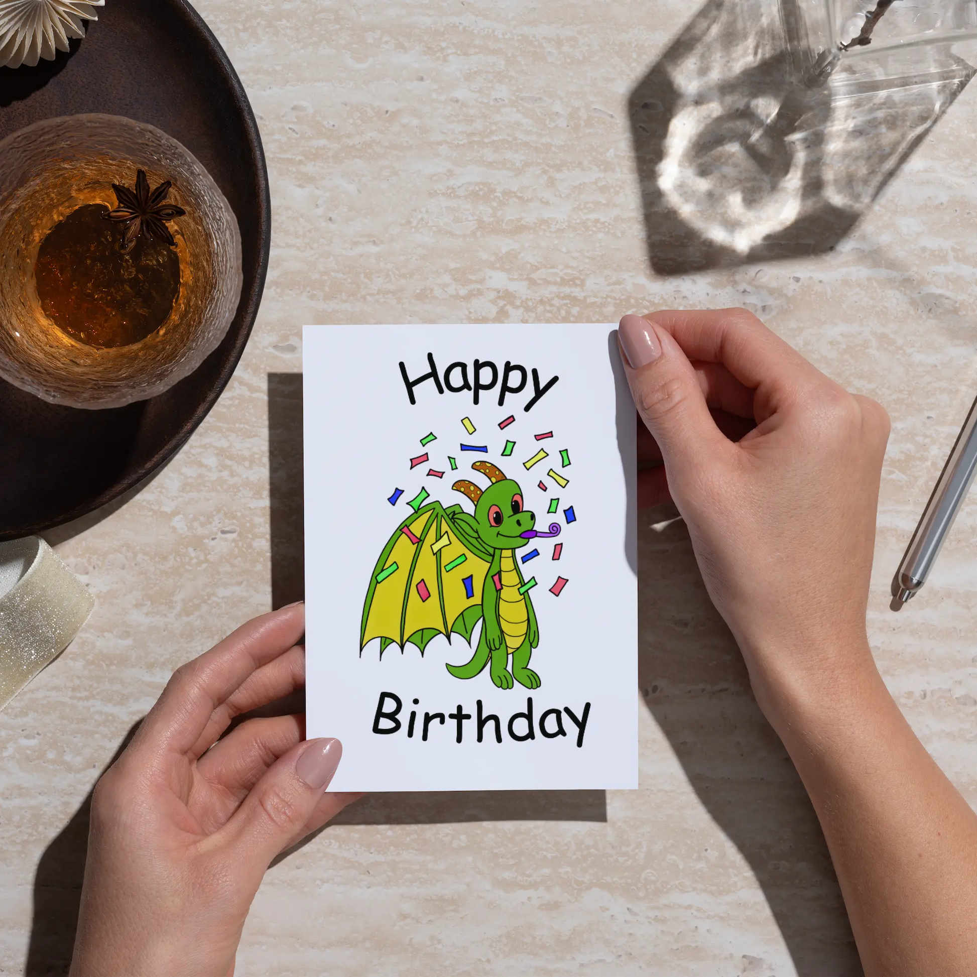 Person holding a birthday card with a green dragon illustration on a table.