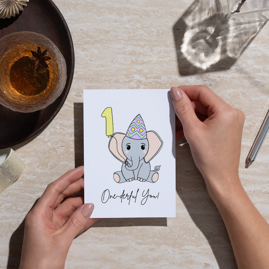 Hand holding a card with an elephant illustration and 'One-derful You!' text on a light surface.