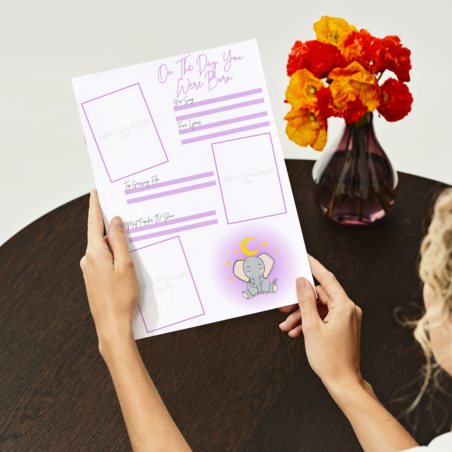 A person holding a printed baby book page with purple and pink text, featuring sections for writing and a space with a moon and stars illustration. A vase with multicolored flowers is visible in the background.